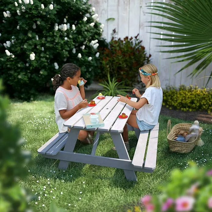 Soulet Kinder-Picknicktisch - L x B x H: 915 x 900 x 500 mm, Holz, Grau - Zwei Mädchen sitzen an einem Kindersitzgarnitur im Garten und spielen mit Spielzeuggeschirr, umgeben von Grün und Blumen.
