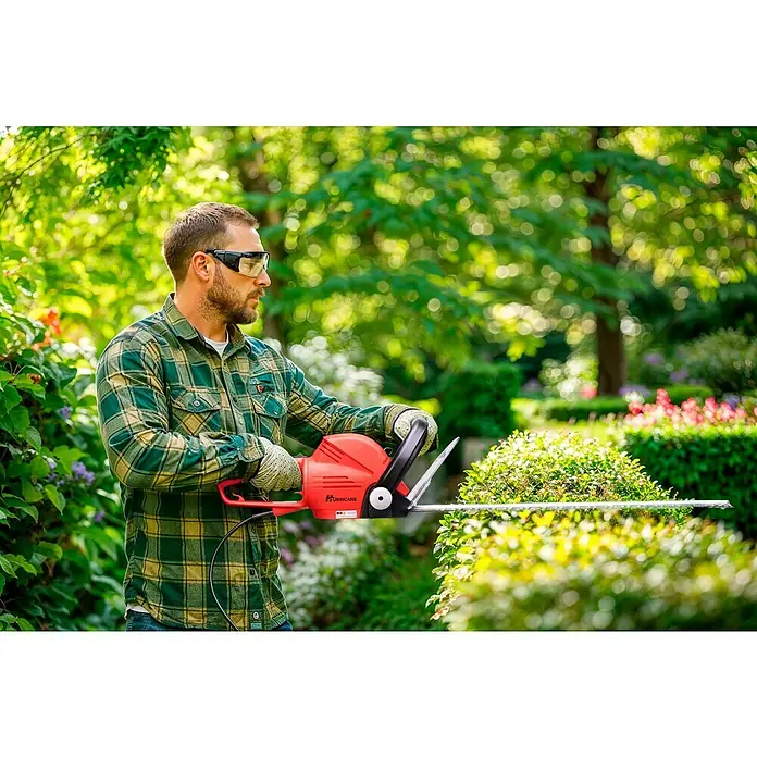 Hurricane Elektro-Heckenschere HHS-I-7560 - 750 W, Schnittlänge: 60 cm, Max. Aststärke: 29 mm - Ein Mann mit Schutzbrille trimmt eine Hecke mit einer roten Hurricane Heckenschere.