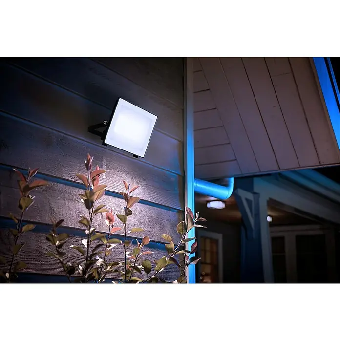 Philips Proyector LED Lois - 24 W, 4.500 lm, 5.000 K, L x An x Al: 20 x 3 x 17 cm, IP65 - Un reflector cuadrado y brillante Philips Hue ilumina una pared de casa, destacando las plantas en primer plano. Iluminación exterior inteligente.