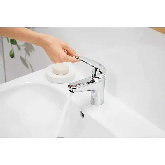 Grohe Grifo Swift XL - Brillante, Cromo - Una mano acciona un elegante grifo cromado mientras el agua fluye hacia un lavabo blanco, junto a un jabón sobre un plato de mármol.