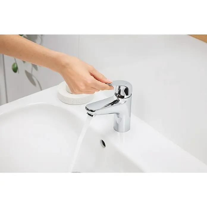 Grohe Grifo Swift XL - Brillante, Cromo - Una mano opera un grifo cromado brillante en un lavabo blanco, con agua corriendo. El grifo moderno es el punto focal en el espacio del baño.