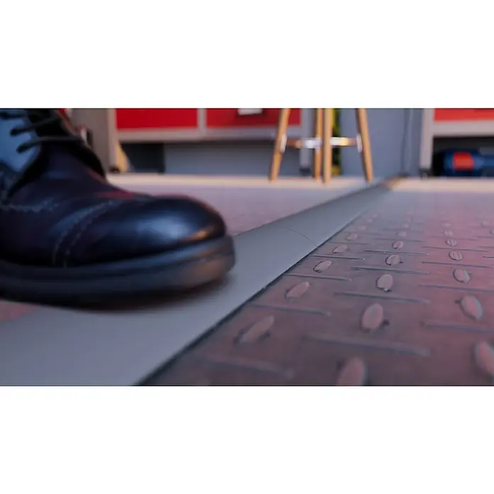 Aufbodenkanal - Länge: 2 m, PVC - Nahaufnahme eines schwarzen Lederschuhs, der auf einem grauen Bodenschild mit Diamantmuster steht. Werkstatt im Hintergrund.