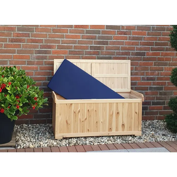 Promadino Gartenbox - L x B x H: 51 x 120 x 56 cm, Holz - Eine hölzerne Gartenbank mit einem blauen Kissen im offenen Stauraum vor einer Backsteinmauer und umgeben von Steinen.