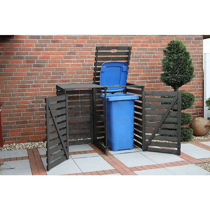 Promadino Mülltonnenbox Variov -  - Mülltonnenbox aus dunklem Holz mit einer blauen Mülltonne darin. Die Box steht auf einem gepflasterten Untergrund vor einer Backsteinmauer.