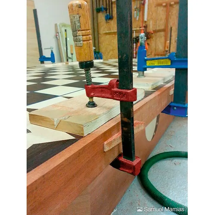 Piher Sargento MM-30 - Anchura de sujeción: 300 mm, Fuerza de tensión: 4.000 N, Apertura máxima: 70 mm - Primer plano de abrazaderas de carpintería que sujetan una pieza de madera unida a una mesa, con una superficie de tablero de ajedrez al fondo.