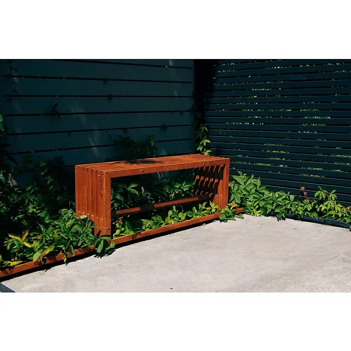 Gartenbank Quadra - B x T x H: 110 x 32,5 x 45 cm, Kiefer, Naturbraun - Rötlich-braune Lamellenholzbank zwischen grünen Pflanzen und dunklen Lattenzäunen auf Beton.
