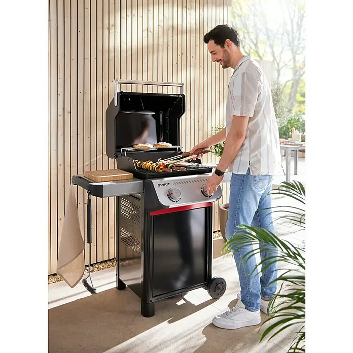 Weber Gasgrill Spirit E-210 - Grillfläche (B x T): 52 x 44 cm, 7,8 kW, Anzahl Brenner: 2 Stk. - Ein Mann grillt auf einem Weber Spirit Grill. Der Grill ist schwarz und silber und hat einen roten Akzent. Es liegt Gemüse und Fleisch auf dem Grill.