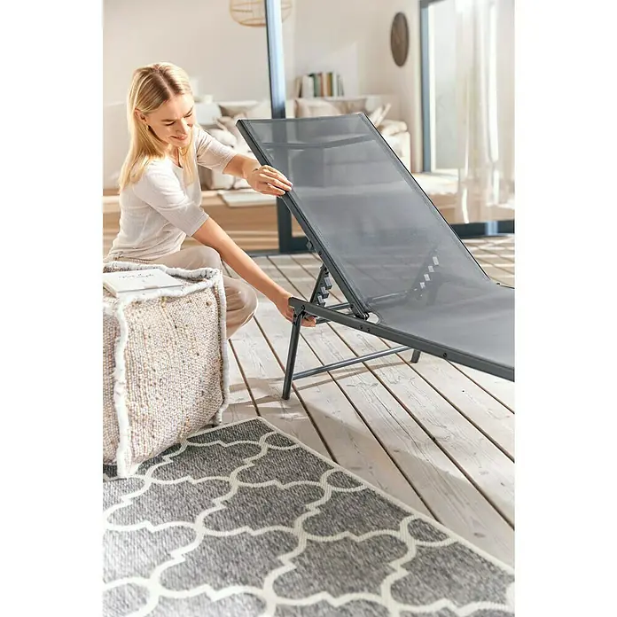 Gartenliege - L x B x H: 181 x 54,5 x 33,5 cm, Dunkelgrau, Stahl, Klappbar - Eine blonde Frau montiert eine graue Sonnenliege auf einer Holzterrasse mit einem Pouf und einem grauen Teppich.