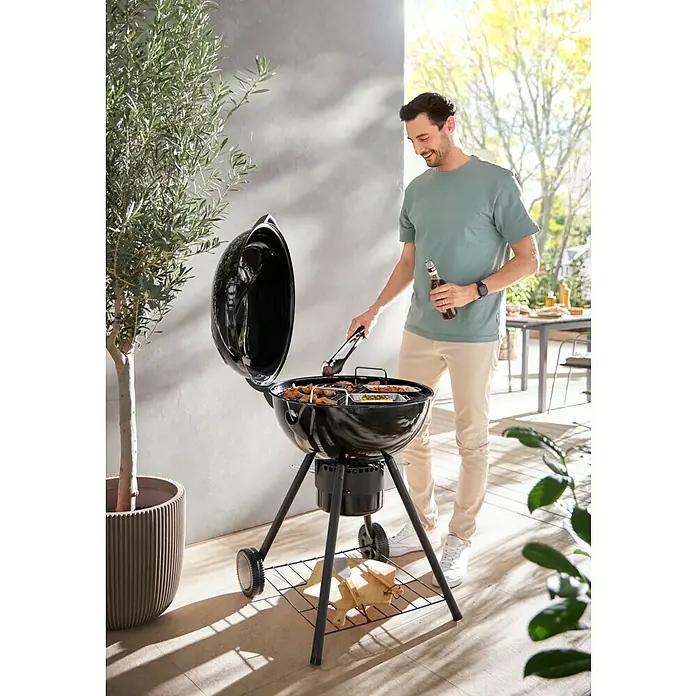 Kingstone Kugelgrill Sunset - Durchmesser Grillfläche: 55 cm, Schwarz - Ein Mann grillt mit einem Holzkohlegrill im Freien. Er verwendet eine Grillzange, um das Essen zu wenden, und hat ein Bier in der Hand.