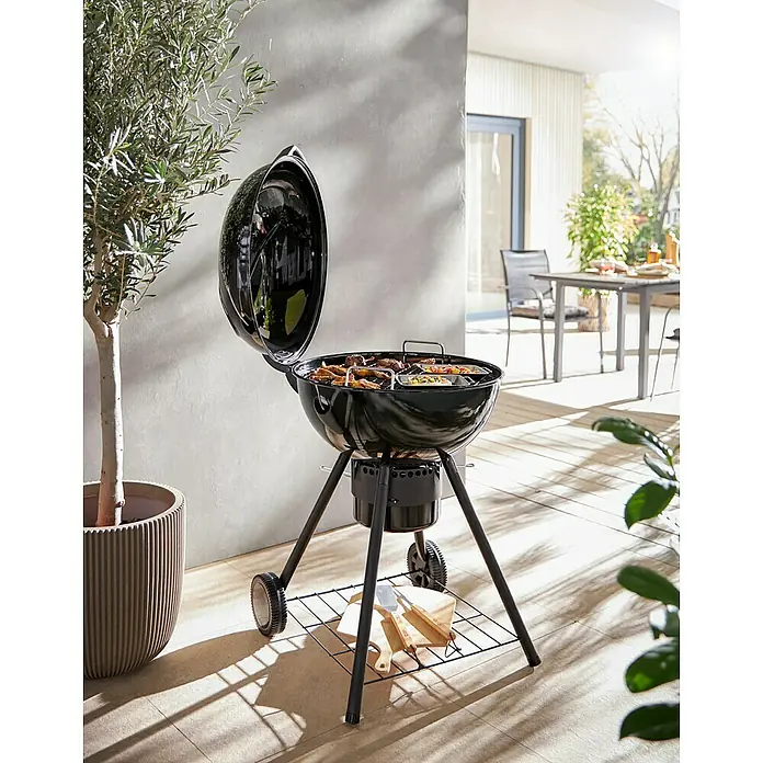 Kingstone Kugelgrill Sunset - Durchmesser Grillfläche: 55 cm, Schwarz - Bild eines schwarzen Kugelgrills mit offenem Deckel auf einer Terrasse, daneben ein Olivenbaum und ein Gartentisch mit Stühlen im Hintergrund.