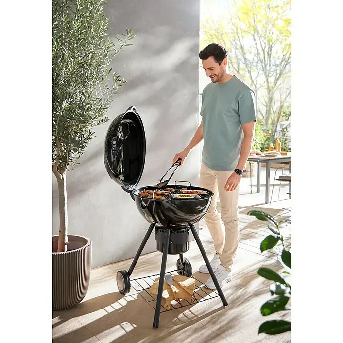 Kingstone Barbacoa redonda con tapa Sunset - Diámetro de la superficie de la parrilla: 55 cm, Negro - Un hombre usa pinzas para voltear comida en una barbacoa de carbón negra en un patio, con leña y herramientas debajo.