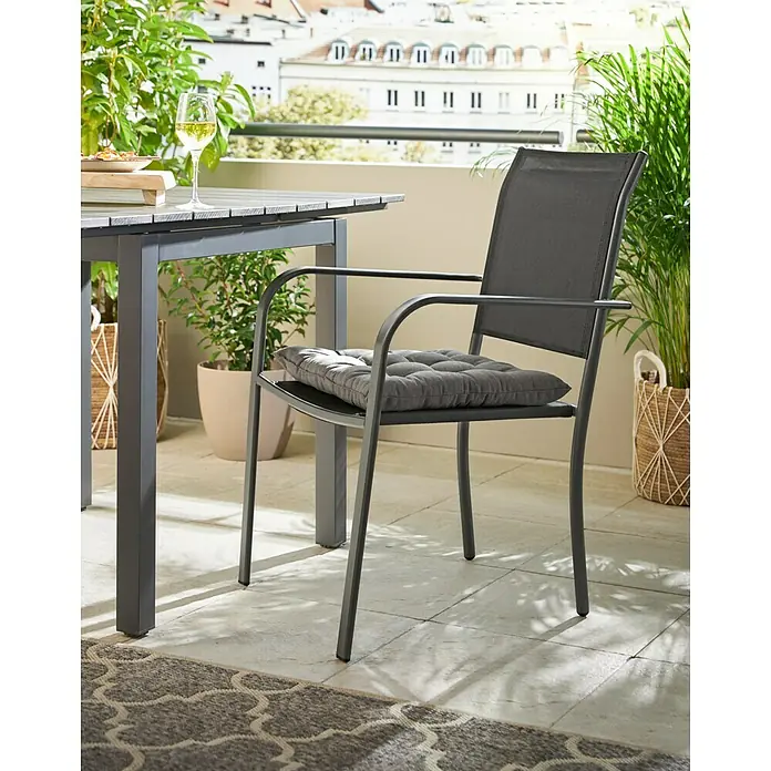 Stapelstuhl - Stahl, Textil, Stapelbar, Night-Grey, B x T x H: 56 x 66 x 86 cm - Nahaufnahme eines grauen Gartenstuhls mit Armlehnen und Sitzkissen auf einer Terrasse mit Pflanzen und Gebäuden im Hintergrund.