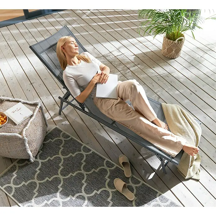 Gartenliege - L x B x H: 181 x 54,5 x 33,5 cm, Dunkelgrau, Stahl, Klappbar - Frau entspannt sich auf einer grauen Sonnenliege im Freien, umgeben von einem Pouf, Teppich und Topfpflanze.