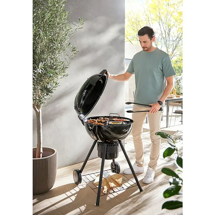 Kingstone Kugelgrill Sunset - Durchmesser Grillfläche: 55 cm, Schwarz - Ein Mann grillt auf einem schwarzen Kugelgrill im Freien. Der Grill ist mit verschiedenen Speisen gefüllt und bereit zum Genießen.