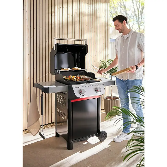 Weber Gasgrill Spirit E-210 - Grillfläche (B x T): 52 x 44 cm, 7,8 kW, Anzahl Brenner: 2 Stk. - Ein Mann grillt mit einem schwarzen Weber Spirit Grill im Freien. Der Grill ist mit Speisen und Grillwerkzeug bestückt.