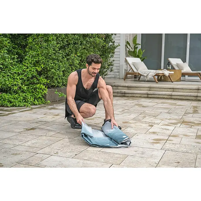 Bestway Lay-Z-Spa Eisbad Glacial Dip - 253 l, Ø x H: 90 x 80 cm - Ein Mann hockt auf Fliesen und rollt eine hellblaue Strandmatte aus. Im Hintergrund sind Liegestühle und Pflanzen zu sehen.