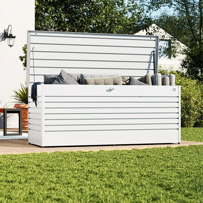 Biohort FreizeitBox Gartenbox 200 - L x B x H: 79 x 201 x 83 cm, Stahl, Weiß - Weiße Lamellen-Außenaufbewahrungsbox, offen und gefüllt mit Kissen und Decke, auf Holzterrasse.