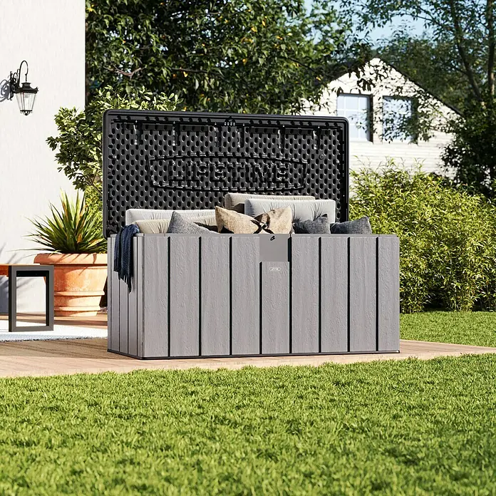 Lifetime Gartenbox - L x B x H: 72 x 151 x 69 cm, Kunststoff, 570 l, Lichtgrau - Geöffnete graue Outdoor-Aufbewahrungsbox auf einer Holzterrasse, gefüllt mit Kissen im Garten.