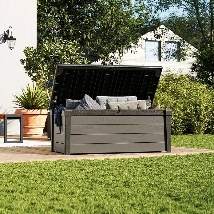 Keter Gartenbox Brushwood - 145 x 69,7 x 60,3 cm, Polypropylen, Taupe - Bild einer geöffneten, grauen Gartenbox aus Kunststoff, die mit Kissen gefüllt ist, im Garten auf einer Holzterrasse.