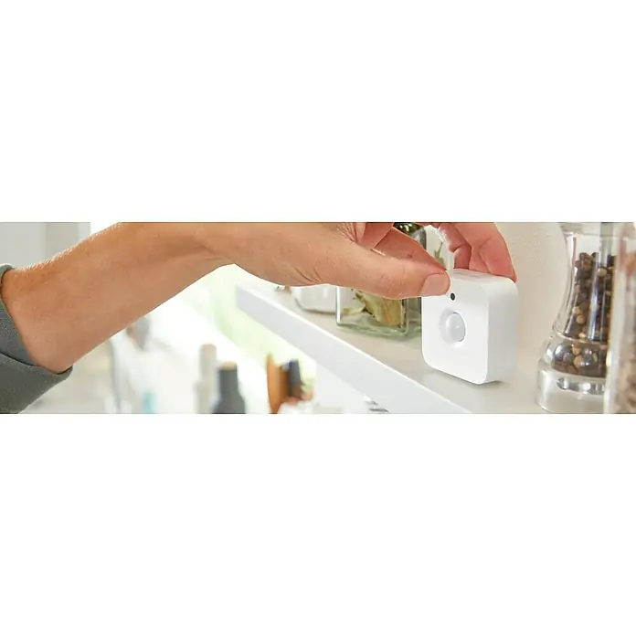 Philips Hue Bewegungsmelder - Weiß, L x B x H: 2 x 5,5 x 5,5 cm - Eine Hand platziert einen Philips Hue Bewegungsmelder auf ein Regal. Es ist ein quadratischer, weißer Sensor zur intelligenten Steuerung von Lampen.