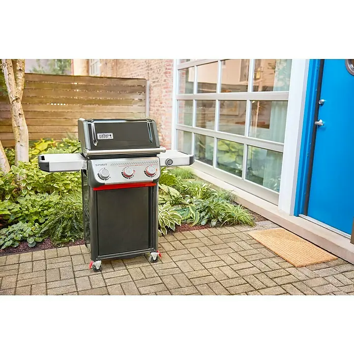 Weber Gasgrill Spirit EP-325 GBS - 9,09 kW, Anzahl Brenner: 3 Stk. - Weber Spirit Gasgrill in einem Hinterhof mit Steinpflaster und üppigem Grün. Der Grill ist schwarz mit silbernen Akzenten.