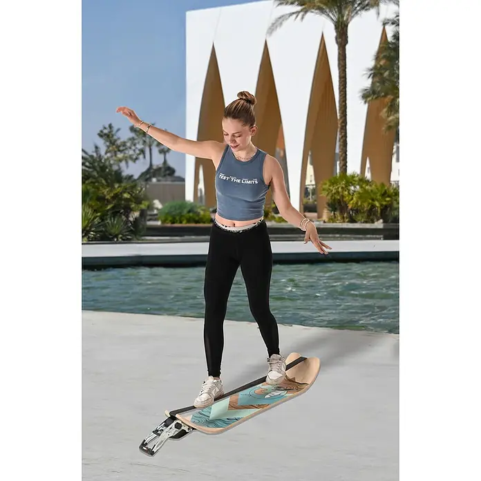 F2 Balance Board Surfline - L x B: 105 x 27,5 cm - Eine junge Frau balanciert auf einem blauen Surfbrett mit dem Aufdruck "Test the Limits" vor einem Gebäude.