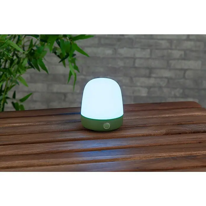 Starlux Akku-Tischleuchte Mini - L x B x H: 10 x 10 x 12 cm, Matcha Green, RGBW, 0,3 W, IP44 - Nahaufnahme einer tragbaren, grünen LED-Lampe mit weißem Lampenschirm auf einem Holztisch vor einer Ziegelwand und Pflanzen.