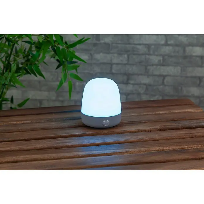 Starlux Akku-Tischleuchte Mini - L x B x H: 10 x 10 x 12 cm, Light Grey, RGBW, 0,3 W, IP44 - Tischleuchte mit weißem Schirm und grauem Fuß auf einem Holztisch, die ein sanftes, warmes Licht für eine entspannte Atmosphäre spendet.