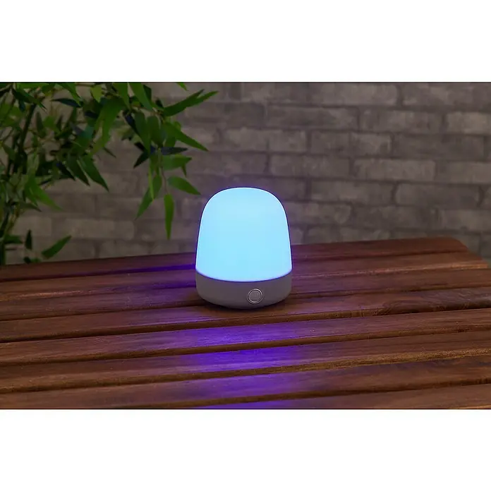 Starlux Akku-Tischleuchte Mini - L x B x H: 10 x 10 x 12 cm, Light Grey, RGBW, 0,3 W, IP44 - Nahaufnahme einer stimmungsvollen blauen Tischlampe, die warmes Licht auf eine hölzerne Oberfläche wirft.