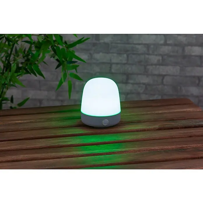 Starlux Akku-Tischleuchte Mini - L x B x H: 10 x 10 x 12 cm, Light Grey, RGBW, 0,3 W, IP44 - Nahaufnahme einer weißen und grauen LED-Tischlampe auf einem Holztisch mit einer grünen Reflexion.
