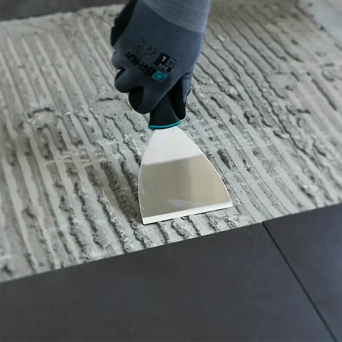 Bihui Schaber-Spachtel TRSC4 - Breite: 100 mm, Edelstahl - Hand in Handschuh verteilt grauen Kleber mit Spachtel für Fliesenverlegung.