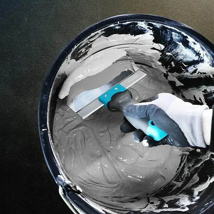 Bihui Eimerkelle PTK165 - Länge: 165 mm - Behandschuhte Hand hält eine Metallkelle mit blauem Griff, mischt grauen Putz in einem schwarzen Eimer.
