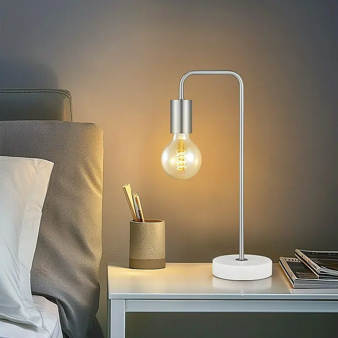 Deko-Tischleuchte Noble - 10 W, L x B x H: 20,3 x 20,3 x 40 cm, Stahl matt - Moderne silberne Tischlampe mit klarer Edison-Glühbirne auf einem weißen Nachttisch im Schlafzimmer.