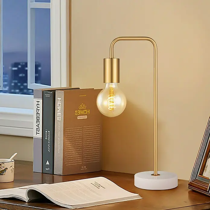 Deko-Tischleuchte Noble - 10 W, L x B x H: 20,3 x 40 x 20,3 cm, Gold - Eine goldfarbene Schreibtischlampe mit freiliegender Edison-Glühbirne und weißem Marmorfuß, die einen Holztisch bei Nacht beleuchtet.