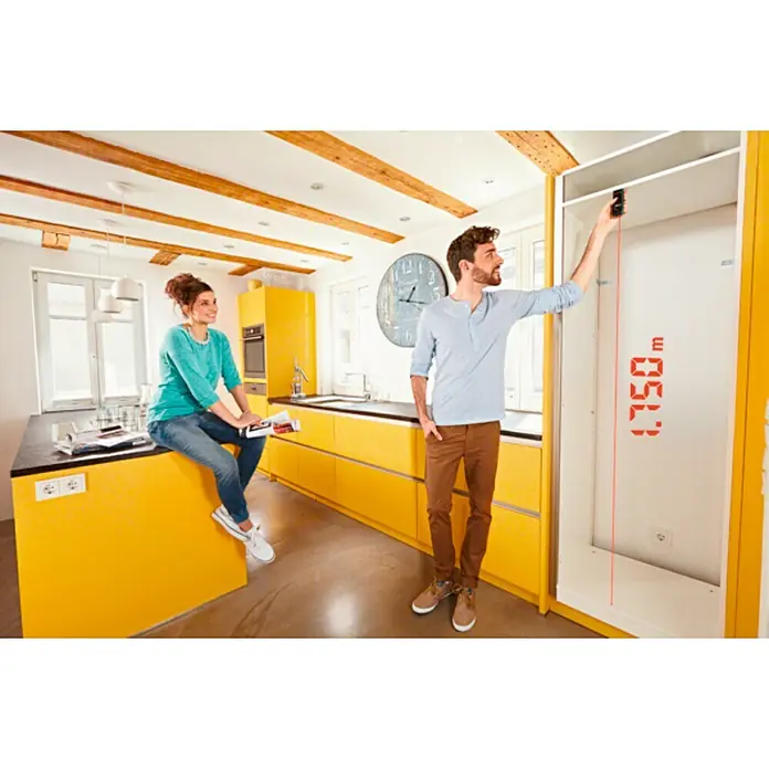 Bosch Medidor de distancia láser de bolsillo Zamo III - Gama de medición: 0,15 m - 20 m - Un hombre mide la altura de un armario en una cocina amarilla con un telémetro láser, mientras que una mujer se sienta en la isla.