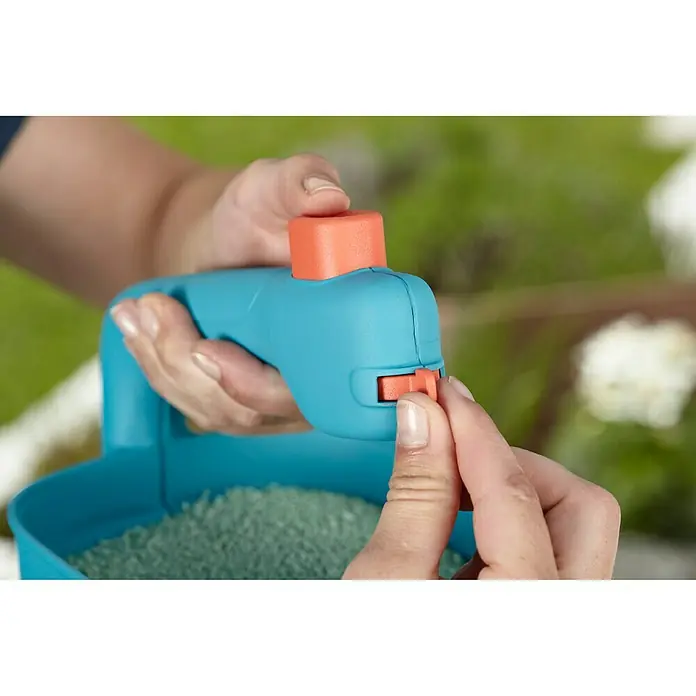 Gardena Handstreuer M - Streubreite: 1 m - 4 m, 1,8 l - Nahaufnahme eines Gardena Streuwagens mit einer Hand, die den orangen Auslöser betätigt, gefüllt mit grünen Samen.