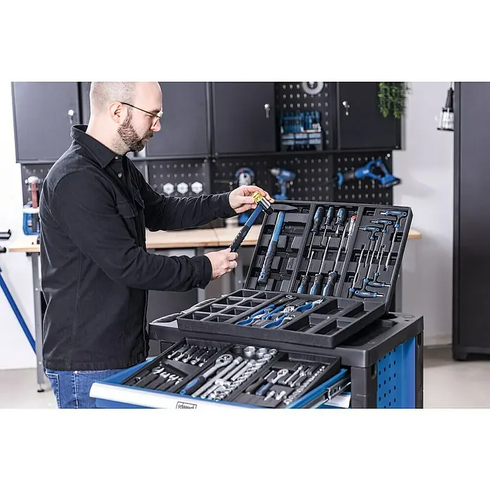Scheppach Carro portaherramientas TW1150 - L x An x Al: 760 x 460 x 965 mm, Número de cajones: 6 ud., 70 pzs. - Un hombre selecciona herramientas de un carro de herramientas Scheppach lleno de una variedad de herramientas, incluidos martillos, destornilladores y alicates.
