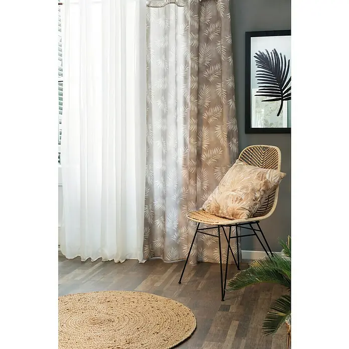 Alfombra de yute Nazar - Beige, Diámetro: 100 cm - Interior acogedor con cortinas blancas translúcidas y cortinas con estampado de hojas junto a una silla de ratán con cojín.