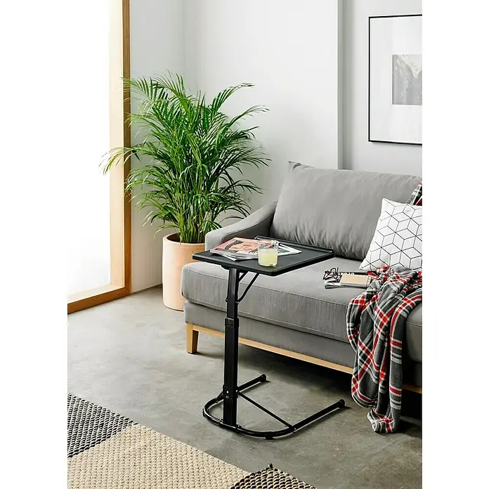 Mesa plegable Multitable - L x An x Al: 43 x 49 x 73 cm, Negro - Una mesa auxiliar negra ajustable se encuentra junto a un sofá gris, adornado con una manta y almohada, ofreciendo comodidad y funcionalidad.