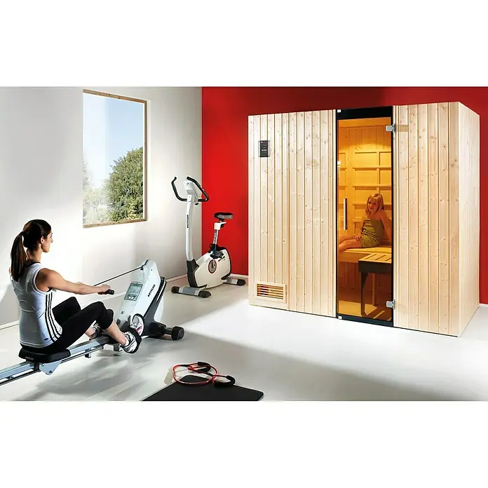Weka Infrarot-Sauna-Kombikabine Uppsala - Therm-Flächenstrahler + 3,6 kW Bio-Saunaofen, 121 x 212 x 199 cm - Ein helles Home Gym mit einem Kettler Rudergerät, einem Heimtrainer und einer Holzsauna mit Glastür und einer Frau darin.
