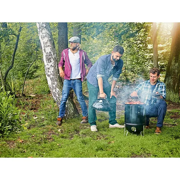 Weber Räucherofen Smokey Mountain CookerDurchmesser Grillfläche: 47 cm, Schwarz - Drei Männer grillen mit einem schwarzen Holzkohlegrill im Wald, während einer das Essen mit einer Zange wendet.
