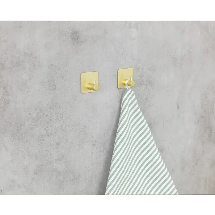 Wenko Colgador de baño Ureo - L x Al: 3 x 4,5 cm, Número de ganchos: 1 ud., Oro - Primer plano de dos ganchos dorados en una pared de hormigón, con una toalla de rayas colgada en uno. Ganchos de pared prácticos.