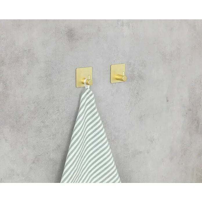 Wenko Colgador de baño Ureo - L x Al: 3 x 4,5 cm, Número de ganchos: 1 ud., Oro - Dos ganchos de pared dorados, uno con una toalla a rayas, contra un fondo gris.
