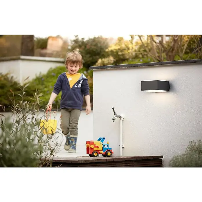 Philips Aplique exterior LED Hegdehog - 42 W, L x An x Al: 16 x 18,2 x 9,1 cm, Antracita, E27 - La elegante luz exterior gris proporciona una luz brillante. Está montada en una pared blanca e ilumina el jardín.