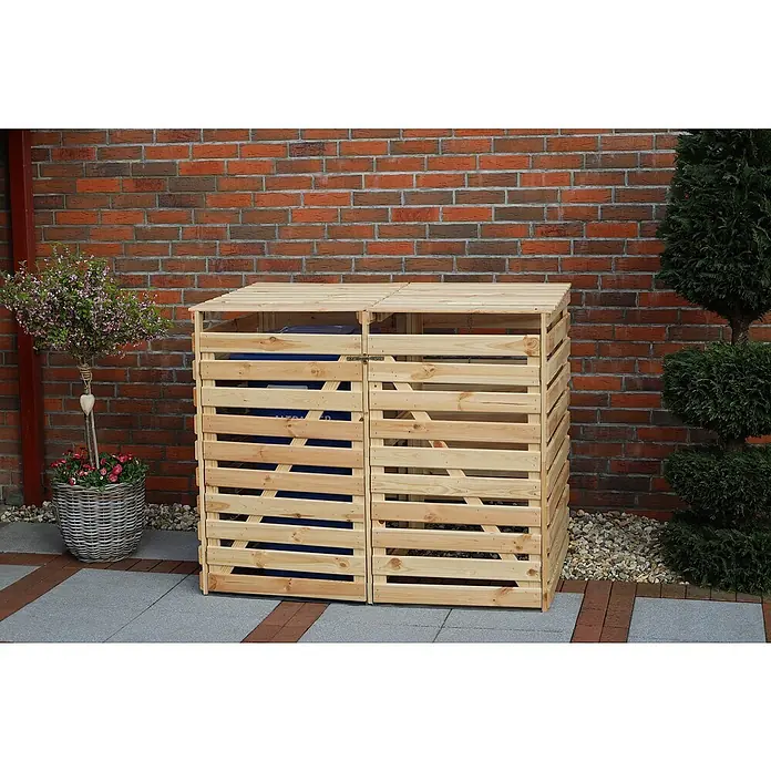 Promadino Mülltonnenbox Vario V -  - Eine hölzerne Doppel-Mülltonnenbox mit Lattenmuster steht vor einer Ziegelmauer und beherbergt blaue Mülltonnen, umgeben von Pflanzen.