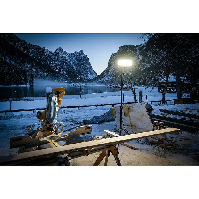 Ledlenser LED Baustrahler AF12C WORK - 8.000 lm, 3.000 K - 6.500 K, 4,2 x 38,2 x 21,4 cm, IP67 - Gelb-silberne Gehrungssäge im verschneiten Bergpanorama im Freien aufgestellt, beleuchtet von einer Arbeitsleuchte, mit Holzbrettern.