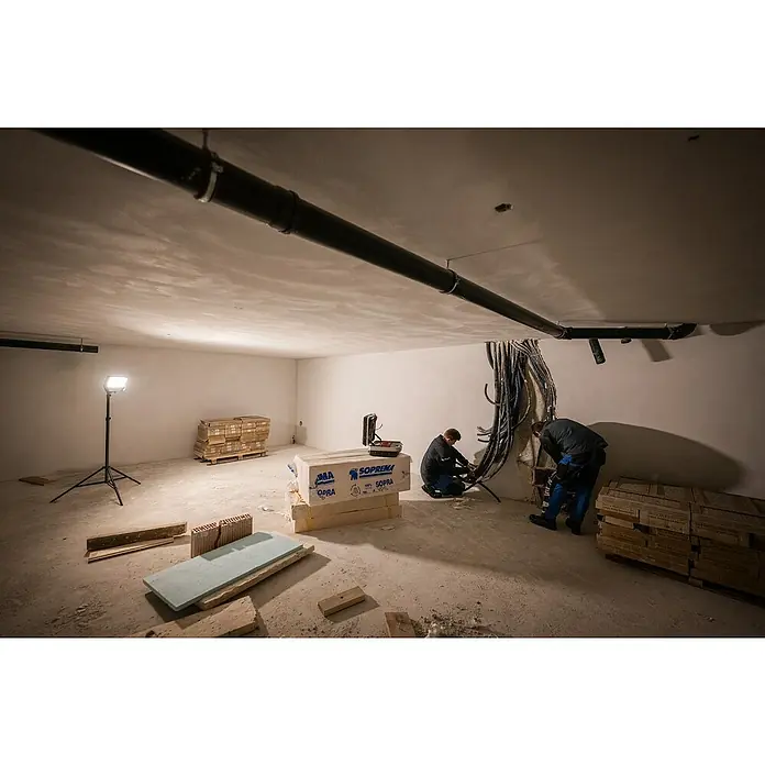 Ledlenser LED Baustrahler AF12R WORK - 8.000 lm, 3.000 K - 6.500 K, 4,1 x 38,2 x 21,4 cm, IP67 - Zwei Arbeiter installieren Kabel in einem Bauraum mit Arbeitsleuchte, Baumaterialien und einer Rohrleitung an der Decke.