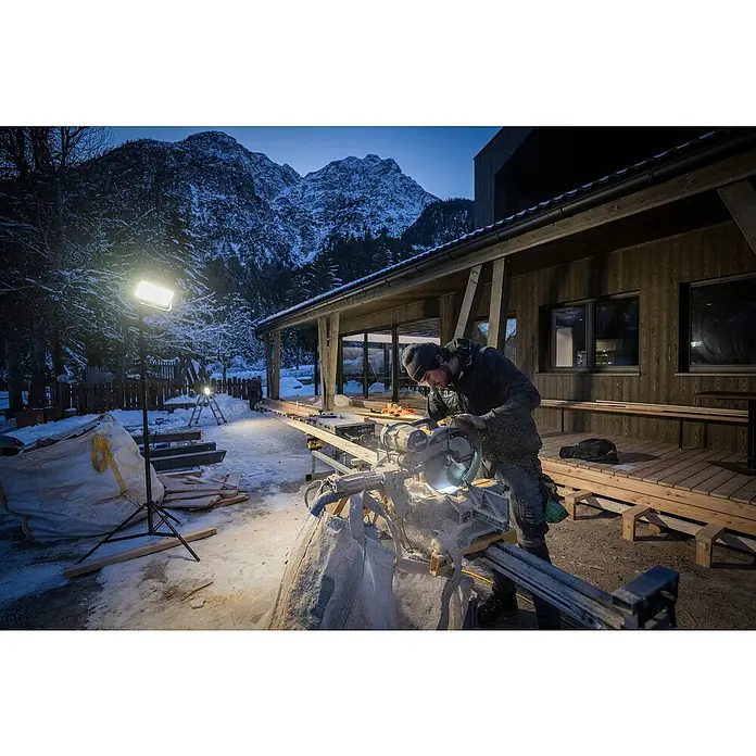 Ledlenser LED Baustrahler AF12R WORK - 8.000 lm, 3.000 K - 6.500 K, 4,1 x 38,2 x 21,4 cm, IP67 - Mann in Winterkleidung schneidet Holz mit einer DeWalt Gehrungssäge im Freien bei Nacht, beleuchtet von Arbeitsleuchten in verschneiter Berglandschaft.