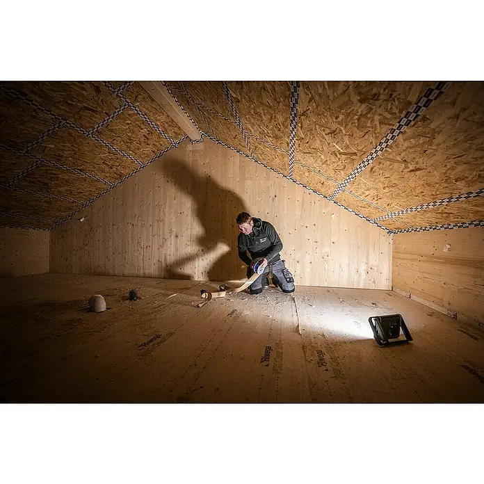 Ledlenser LED Baustrahler AF8R WORK - 4.000 lm, 3.000 K - 6.500 K, 4,1 x 27,5 x 17,9 cm, IP67 - Arbeiter verklebt hellbraunes Dichtband an Isolationsfugen im Holzdachboden.