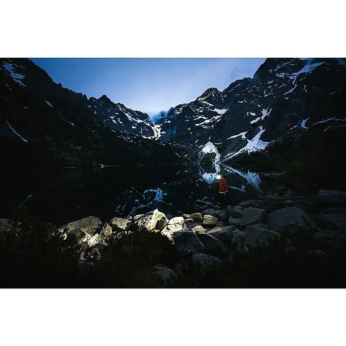 Ledlenser Stirnlampe HF8R Core - 1.600 lm, IP68 - Person in roter Jacke mit Stirnlampe, die Felsen an einem dunklen Bergsee beleuchtet.